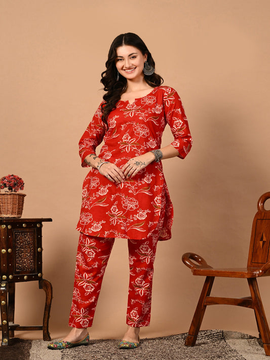 Woman wearing a red floral outfit standing against a beige wall with a wooden chair and table.