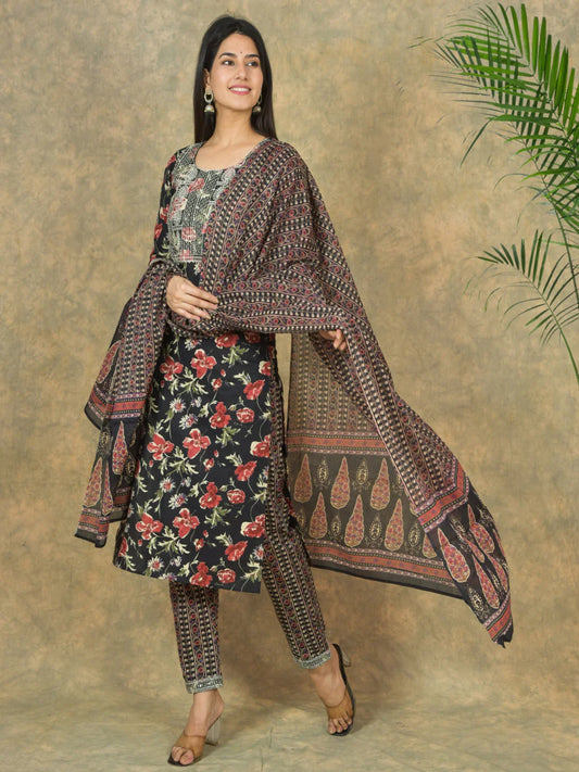 Woman wearing a traditional outfit with a floral dress and patterned shawl against a textured wall.