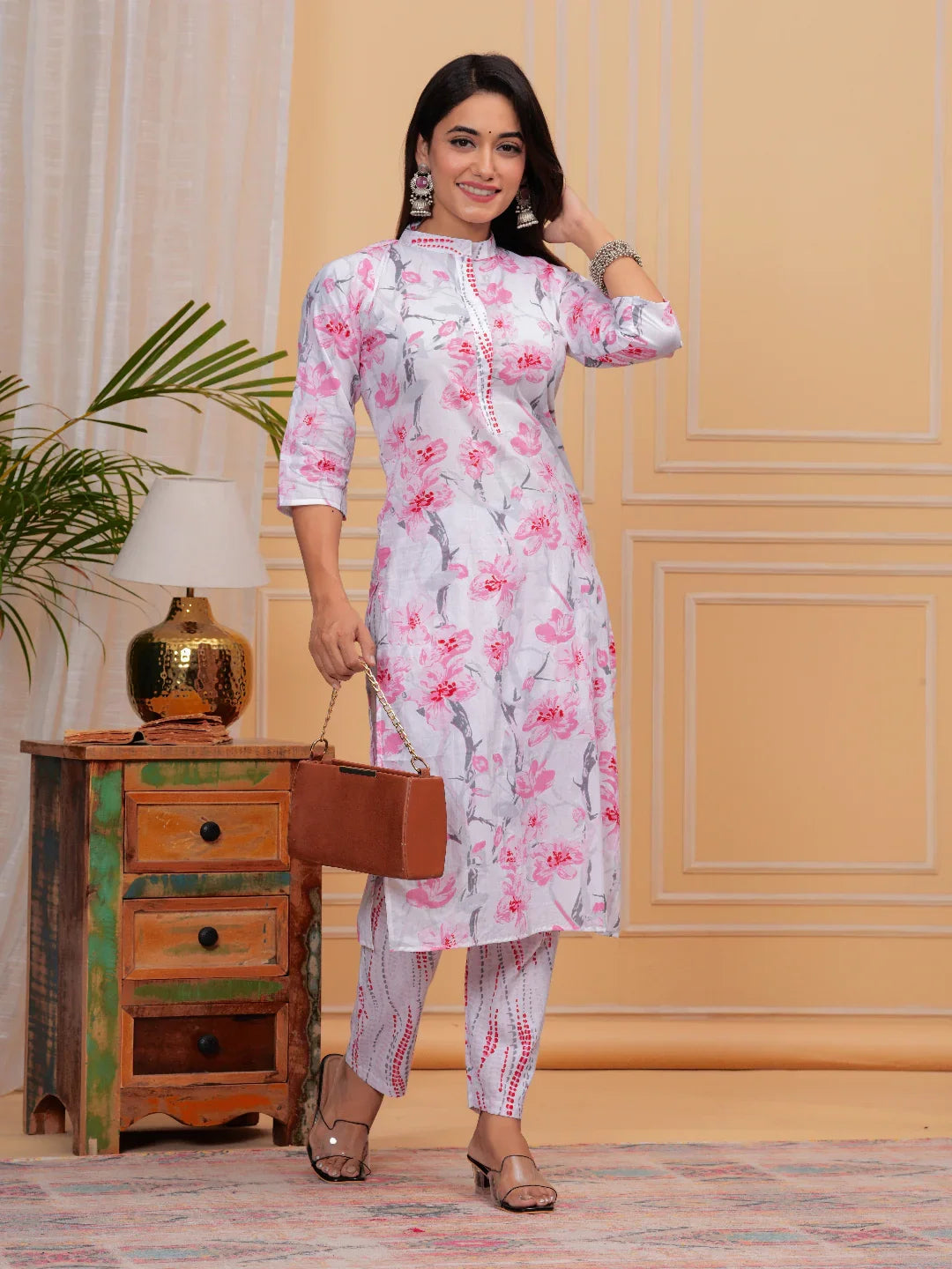 Woman in a floral dress holding a suitcase in a room with a lamp and plant.