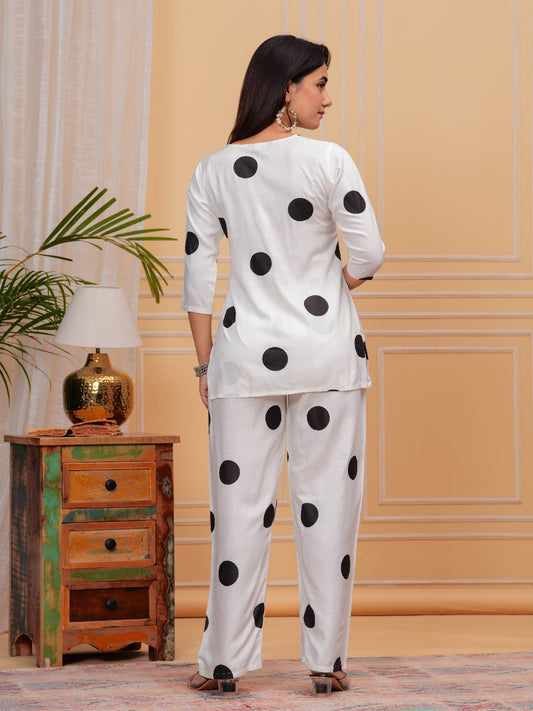 Woman wearing a white outfit with black polka dots in a room with a wooden cabinet and lamp.