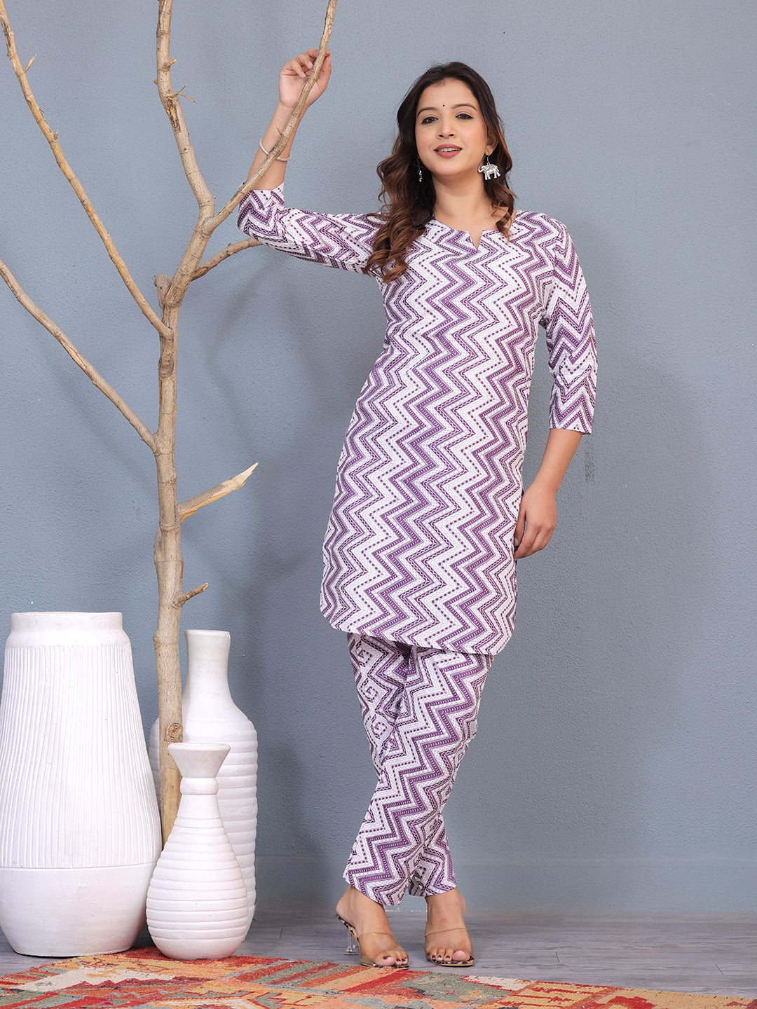 Woman wearing a purple and white zigzag patterned outfit standing next to decorative vases against a gray background. > Mitrika