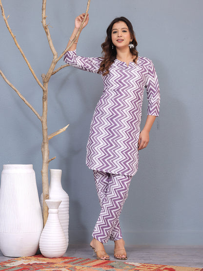 Woman wearing a purple and white zigzag patterned outfit standing next to decorative vases against a gray background. > Mitrika