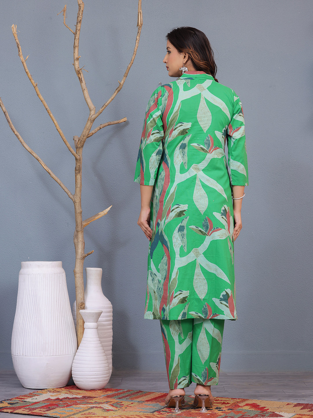 Woman wearing a green floral outfit standing in a room with white vases and a gray wall. > Mitrika
