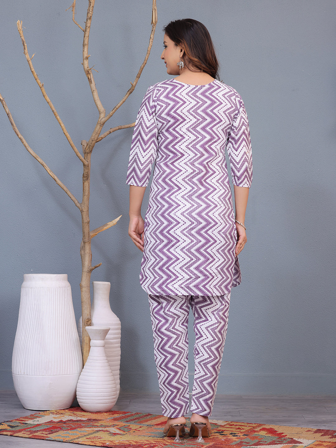 Woman wearing a purple and white zigzag patterned outfit standing against a gray wall with decorative vases. > Mitrika