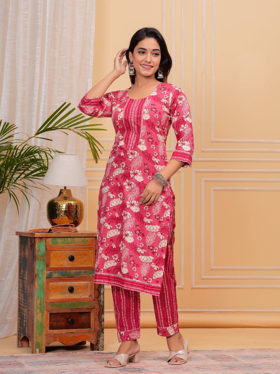 Woman wearing a pink floral kurta with pants in a room with a lamp and plant.