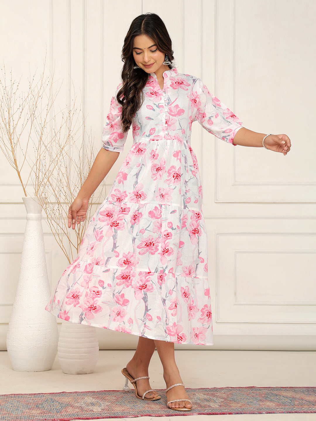 Woman wearing a floral dress standing in a room with a white wall and decorative rug. > Mitrika