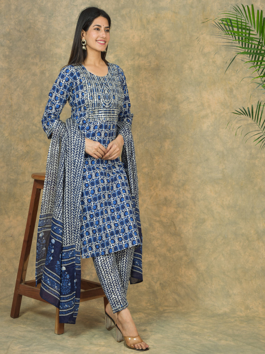 Woman wearing a blue and white traditional outfit standing next to a wooden stool against a textured wall.
