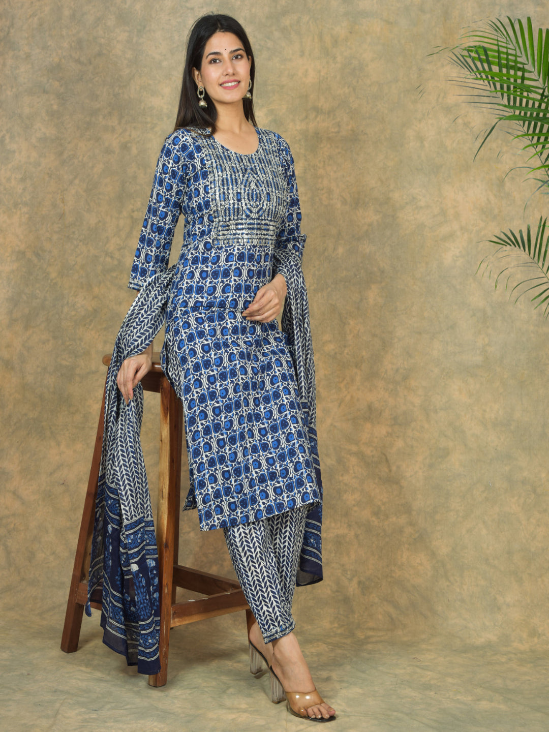 Woman wearing a blue and white patterned outfit sitting on a wooden stool against a textured wall.