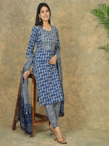 Woman wearing a blue and white patterned outfit sitting on a wooden stool against a textured wall.