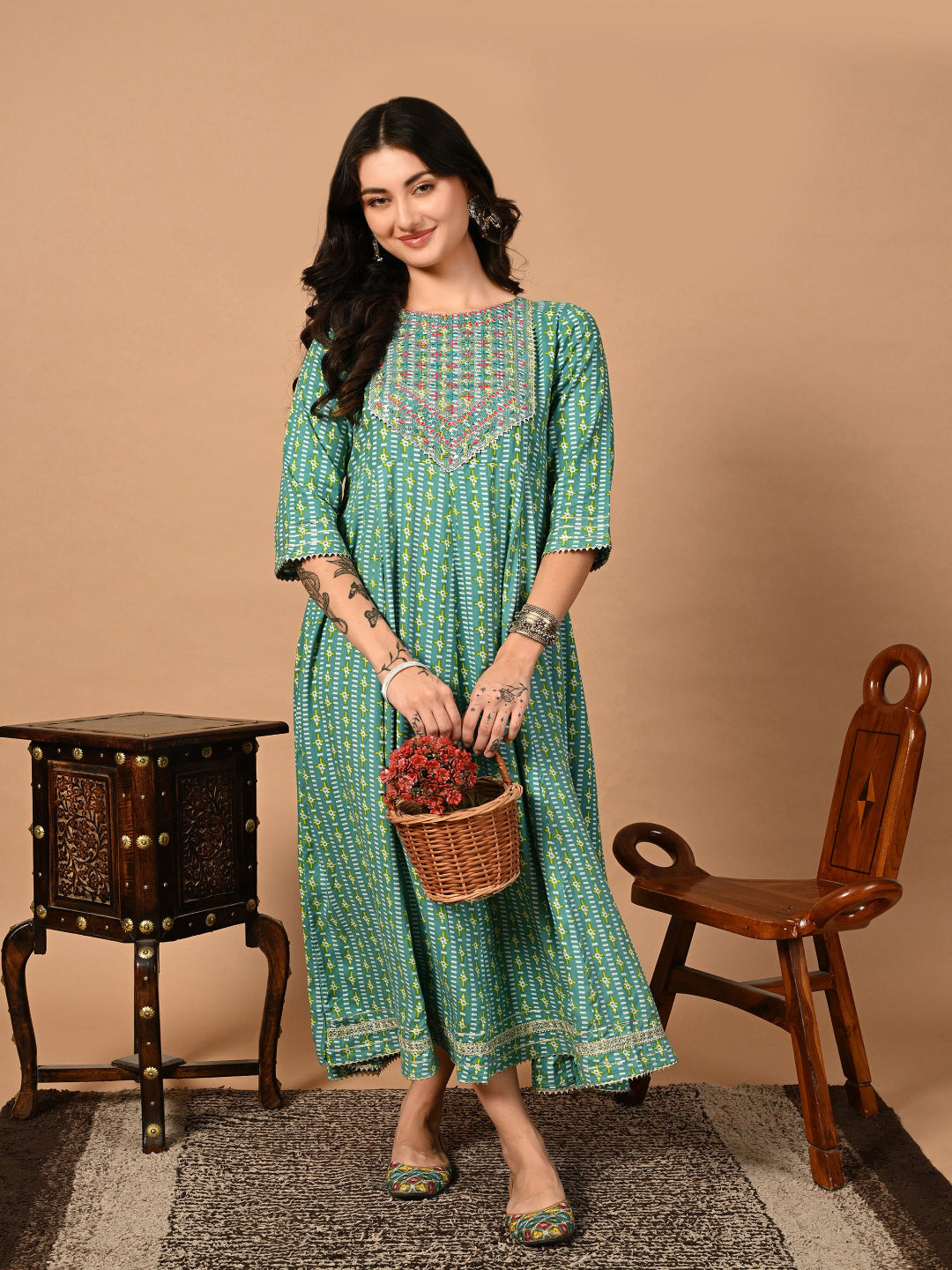 Woman in a green patterned dress holding a basket with flowers against a beige background >Mitrika