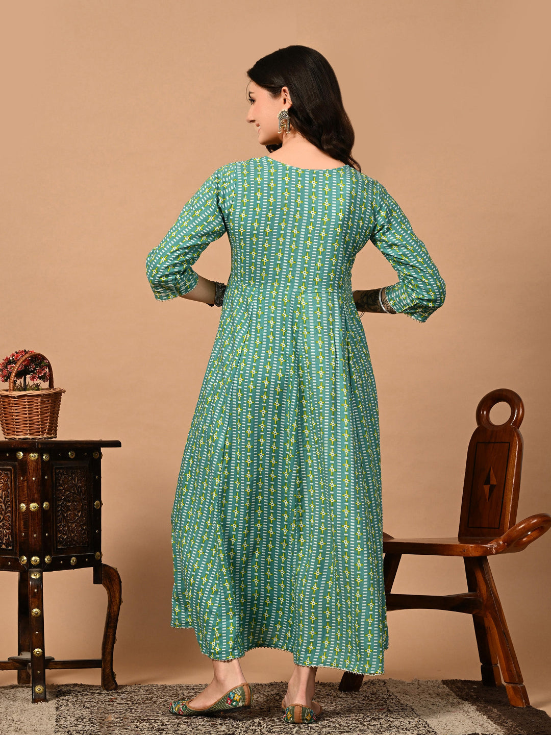 Woman wearing a green patterned dress standing in front of a beige wall with a wooden chair and table.