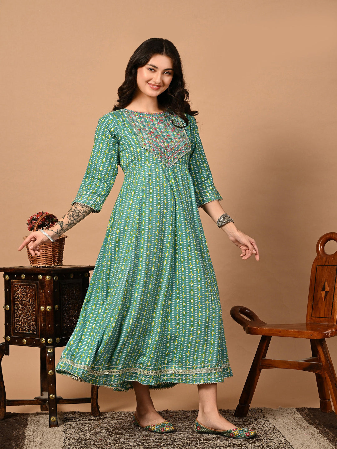 Woman wearing a green patterned dress standing in front of a wooden chair and table.