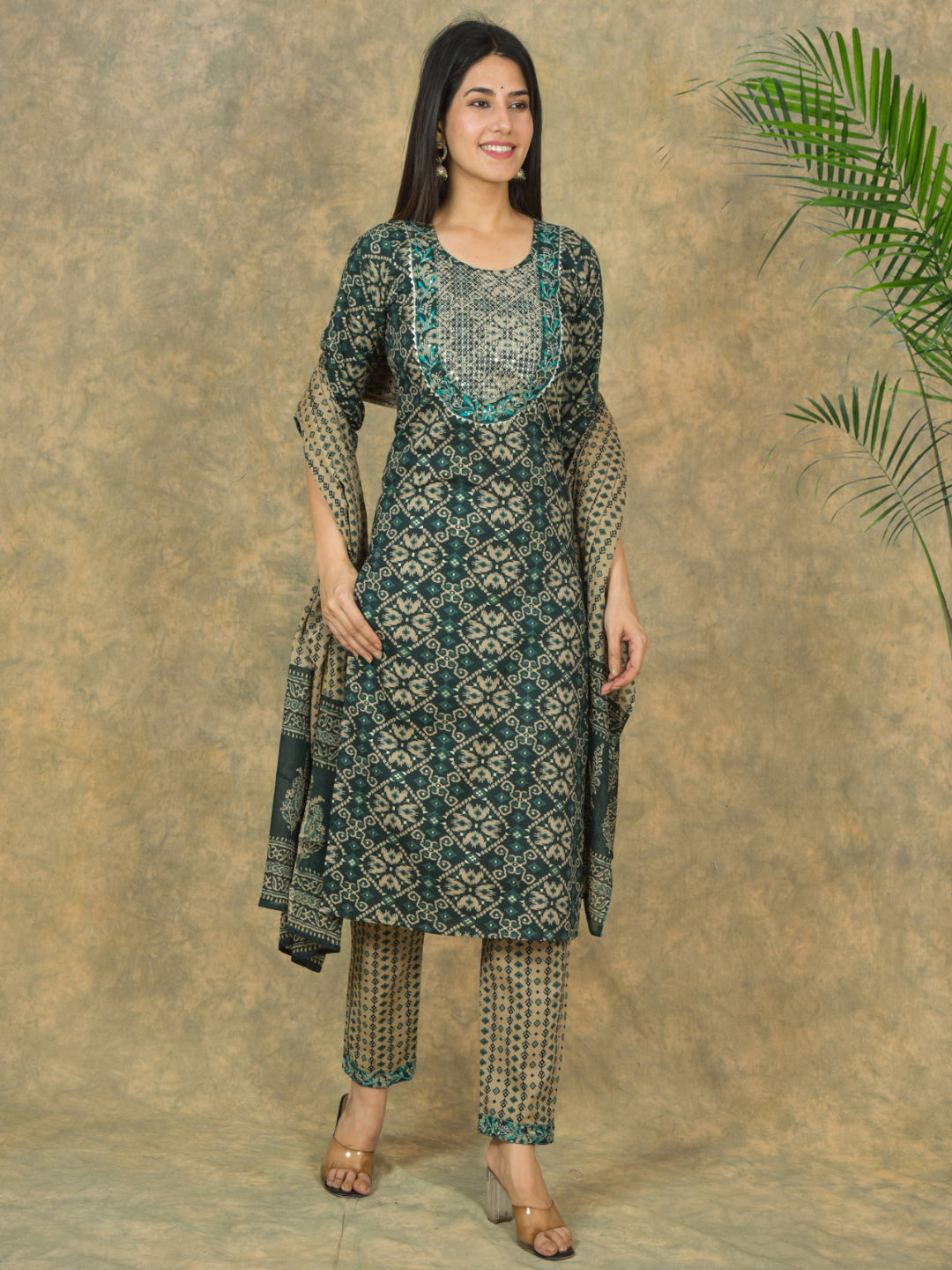 Woman wearing a green and black patterned kurta with pants against a textured brown background.