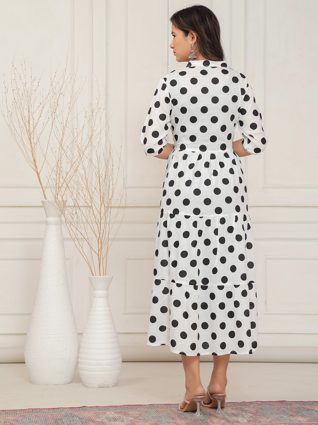 Woman wearing a polka dot dress standing in a room with white walls and decorative vases.