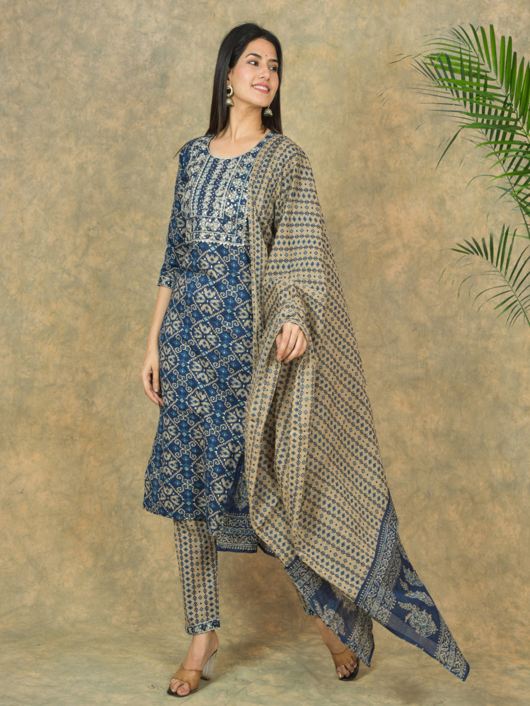 Woman wearing a blue and beige traditional outfit with a patterned dupatta against a textured beige background.