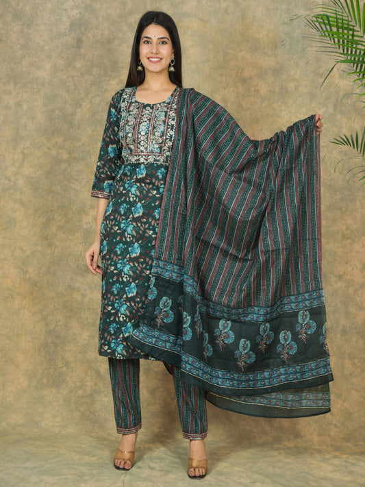 Woman wearing a traditional outfit with a floral top, striped dupatta, and matching pants against a textured wall. > Mitrika