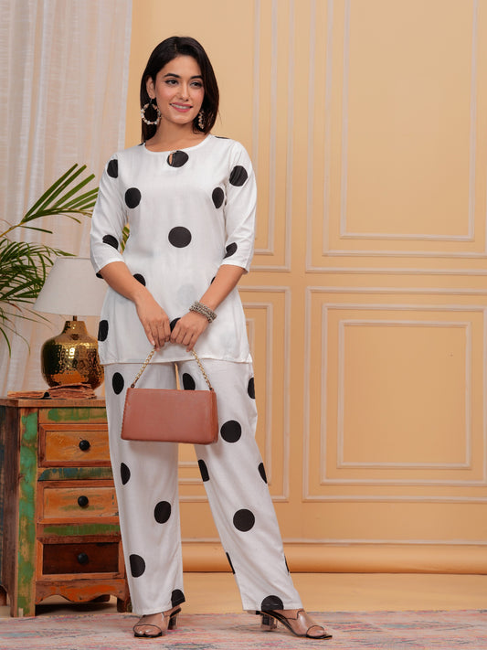 Woman in a white outfit with black polka dots standing in a room with decorative elements. > Mitrika