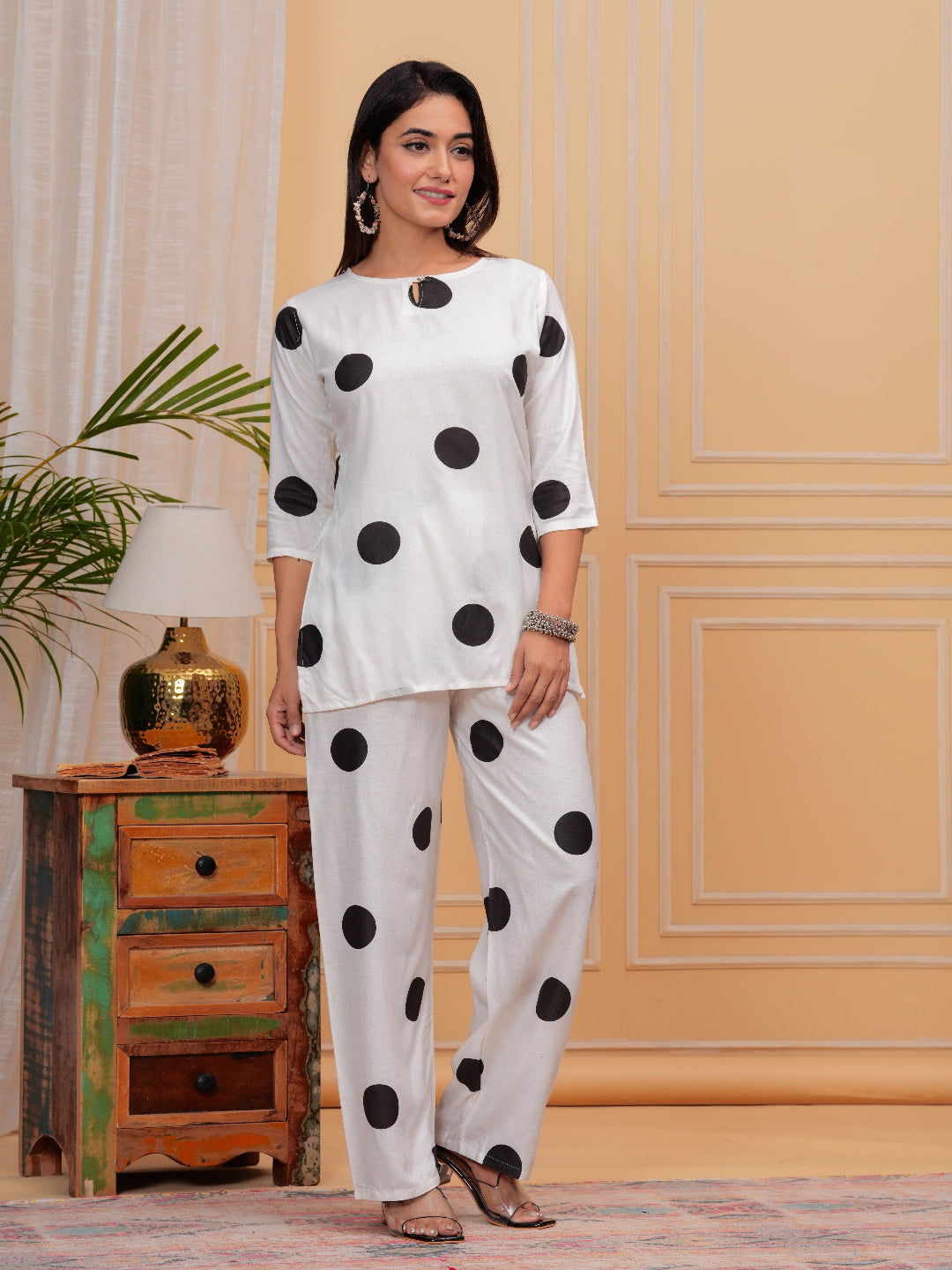 Woman wearing a white outfit with black polka dots in a room with a wooden cabinet and plant.