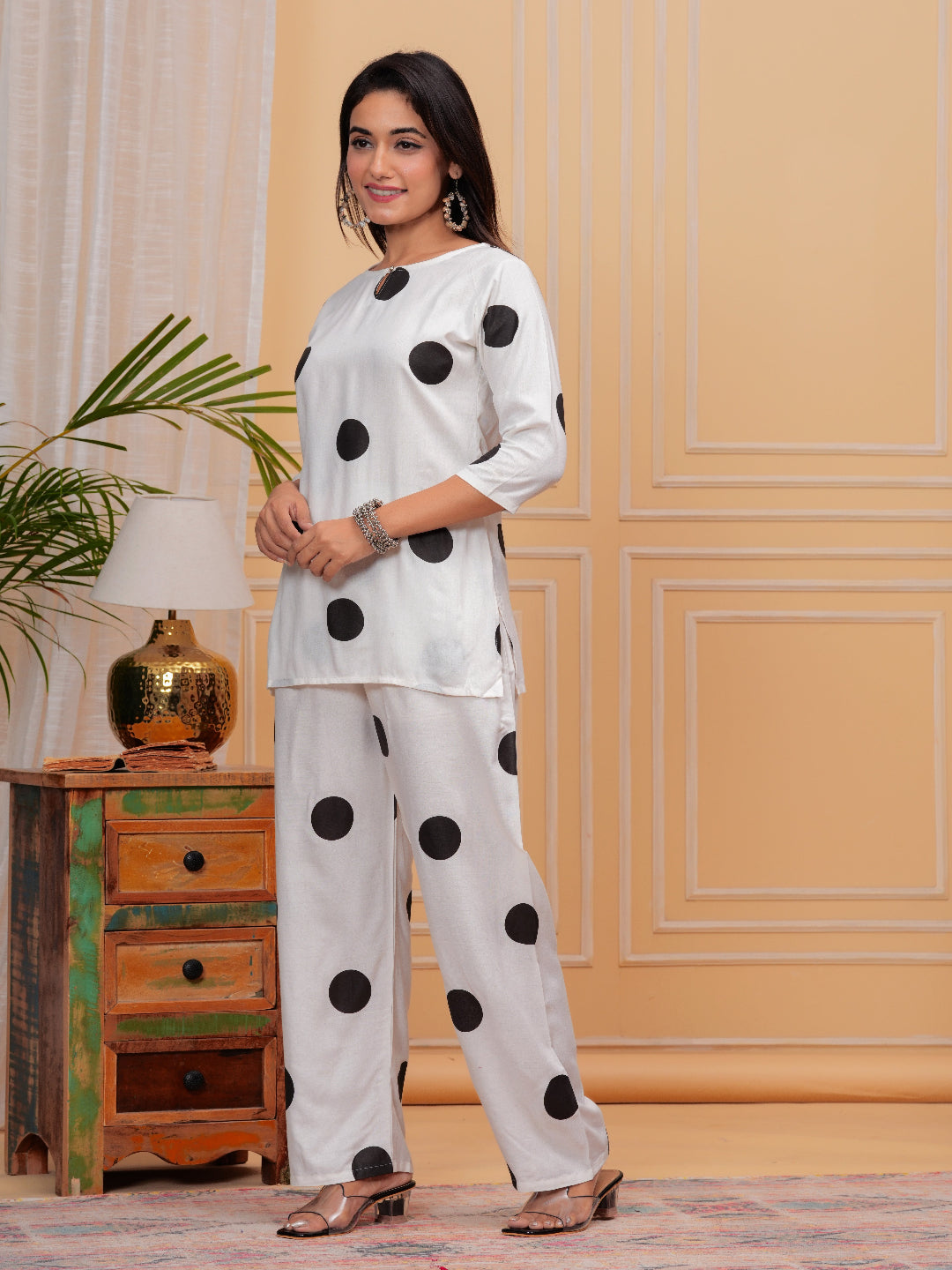 Woman wearing a white outfit with black polka dots in a room with a wooden cabinet and lamp.