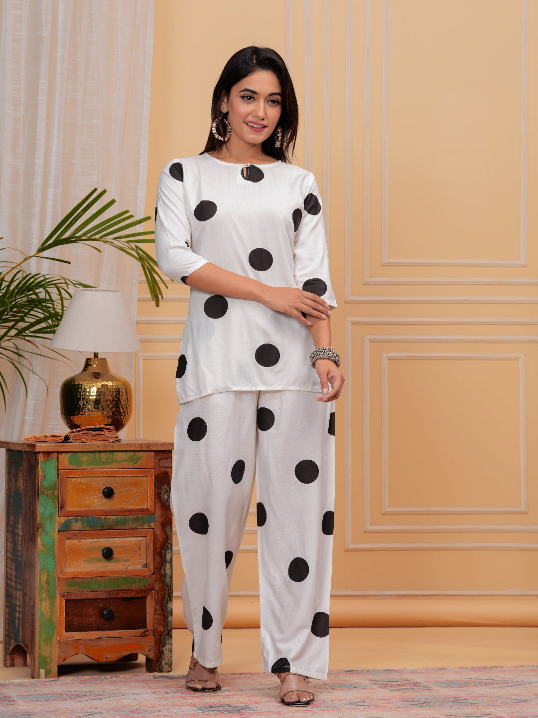 Woman wearing a white outfit with black polka dots standing in a room with a wooden cabinet and lamp.