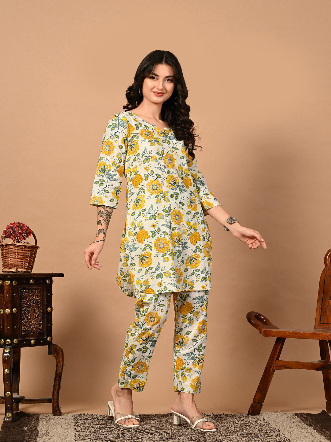 Woman wearing a floral outfit standing against a beige wall with a wooden chair and table in the background.